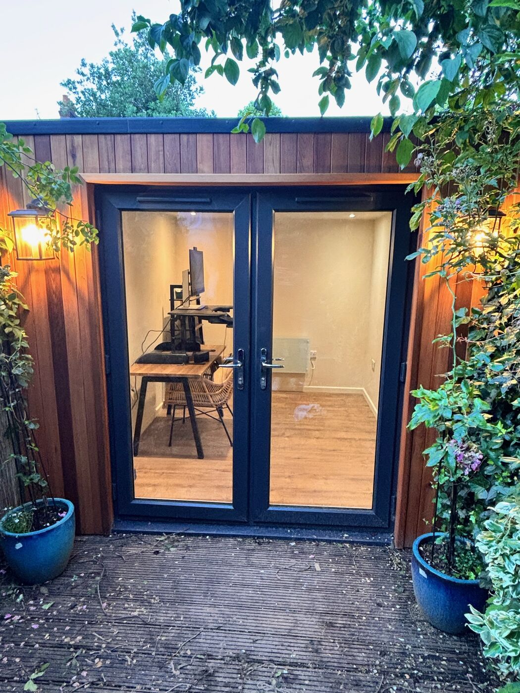 Cedar garden office with double doors and wall lantern, set among climbing plants