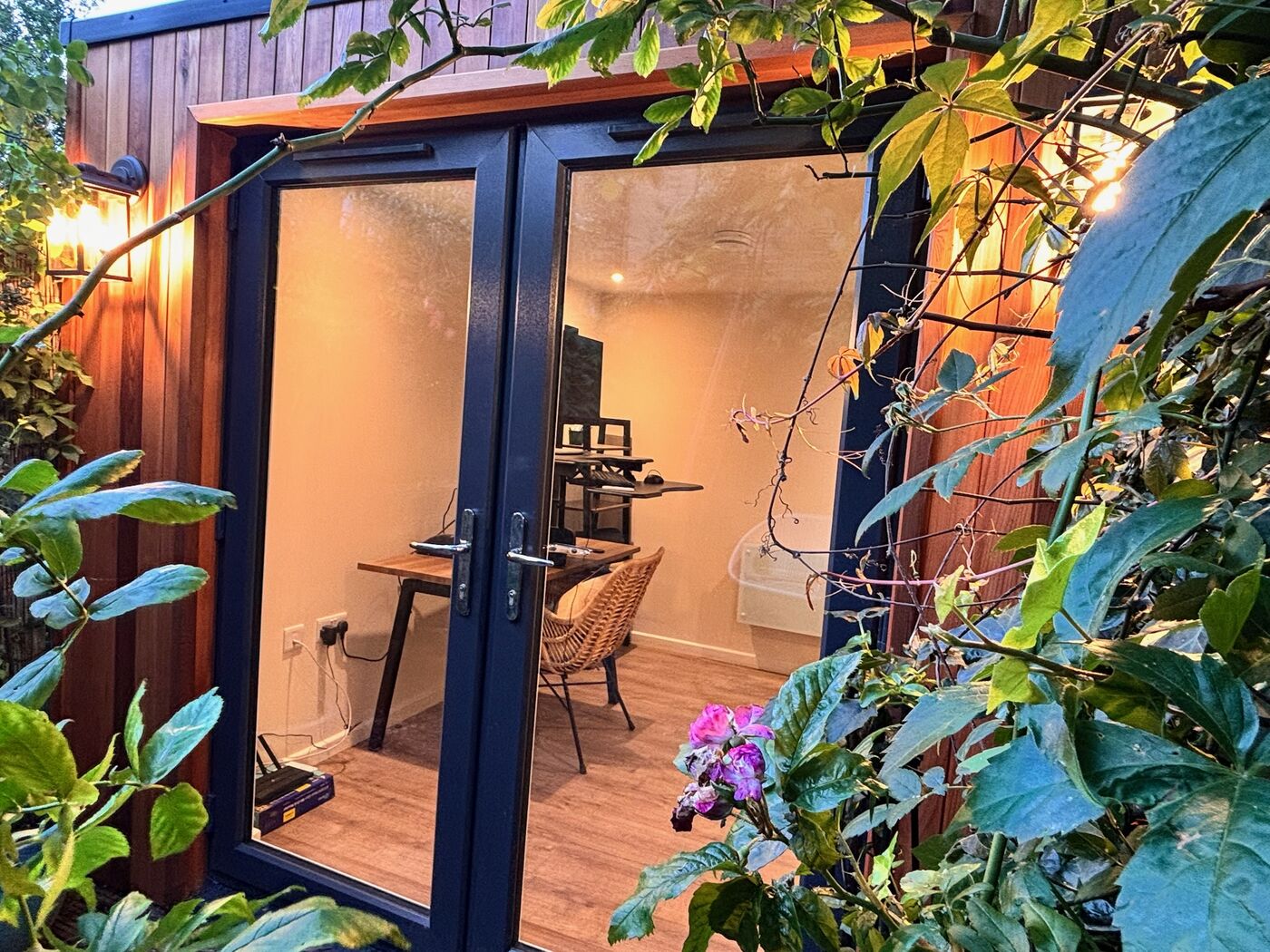 Garden office doors seen through foliage, with a desk and chair inside